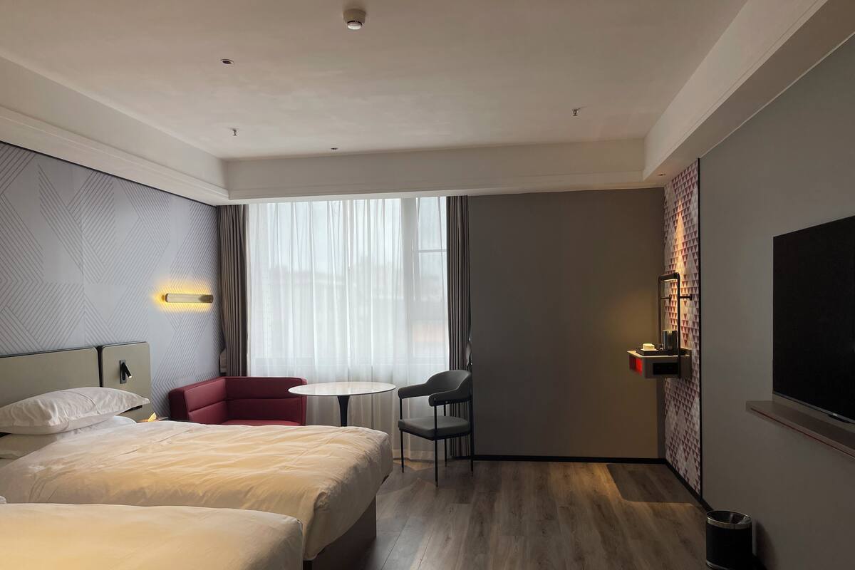 A hotel room with two beds, a red armchair, a small round table, a TV, and a window with sheer curtains.