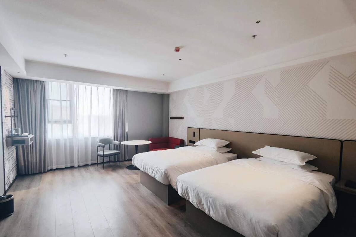 A hotel room with two beds, a small table, a chair, and a large window with curtains.