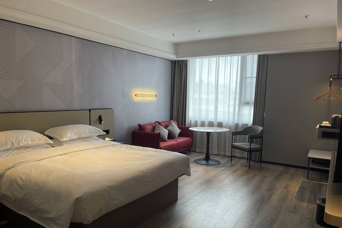 A hotel room with a large bed, a red sofa, a small round table, and a chair.