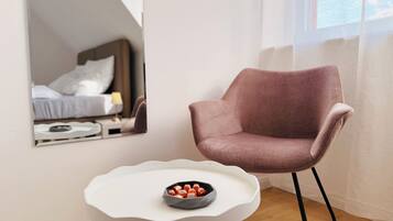 Ein modernes Zimmer mit einem weißen Stuhl, einem kleinen weißen Tisch mit einer Schale mit Snacks und einem großen Spiegel, der ein Bett und einen Nachttisch reflektiert.