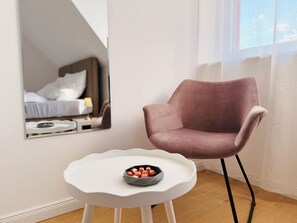 A modern room with a white chair, a small white table with a bowl of snacks, and a large mirror reflecting a bed and a nightstand.
