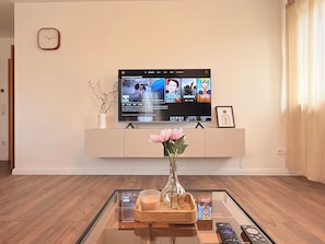 Un saló amb una televisió de pantalla plana sobre un moble blanc, un vas amb flors damunt una taula de café de vidre, i un rellotge a la paret.