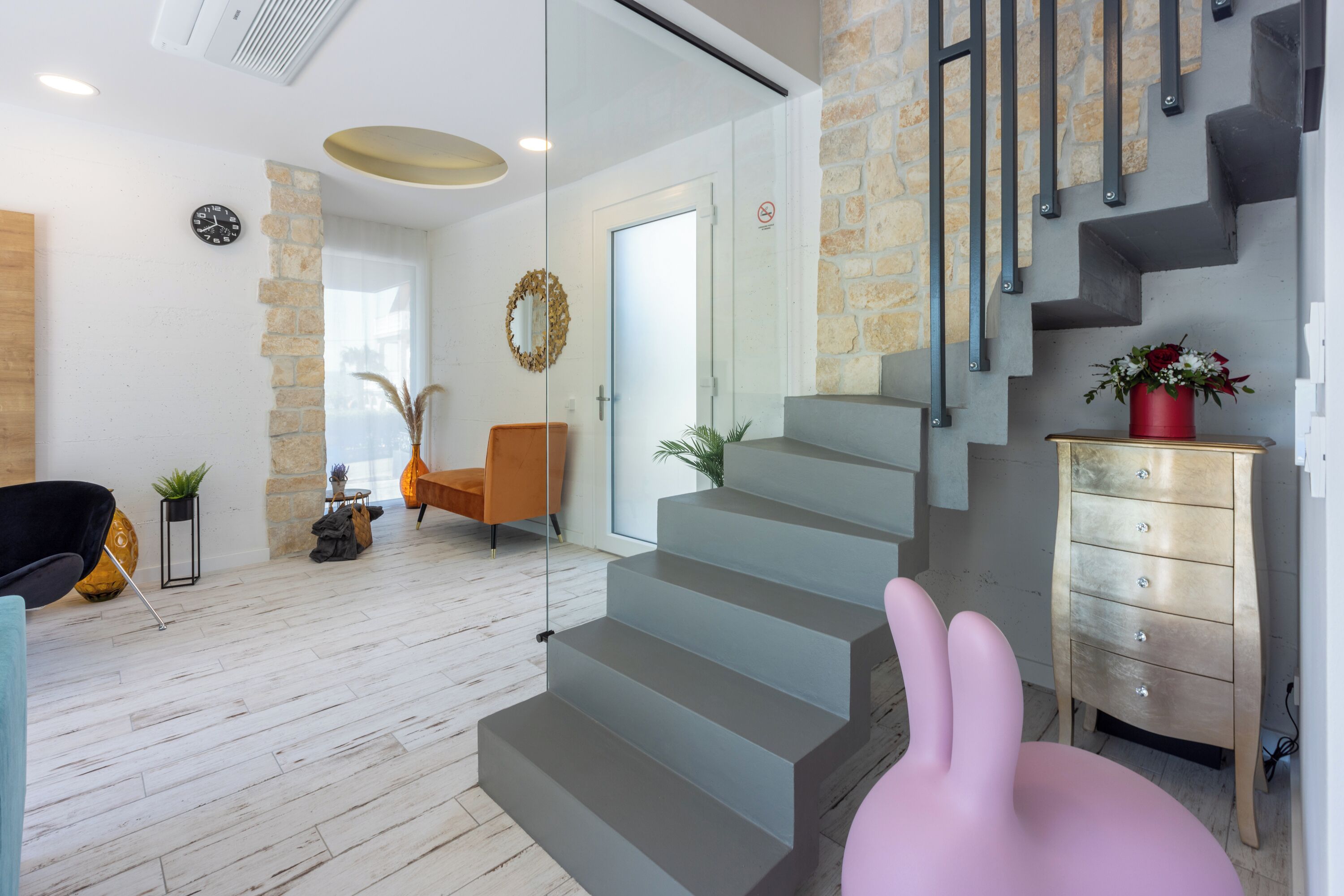 A modern interior with a staircase, a wooden floor, a chair, a dresser, and a wall clock.