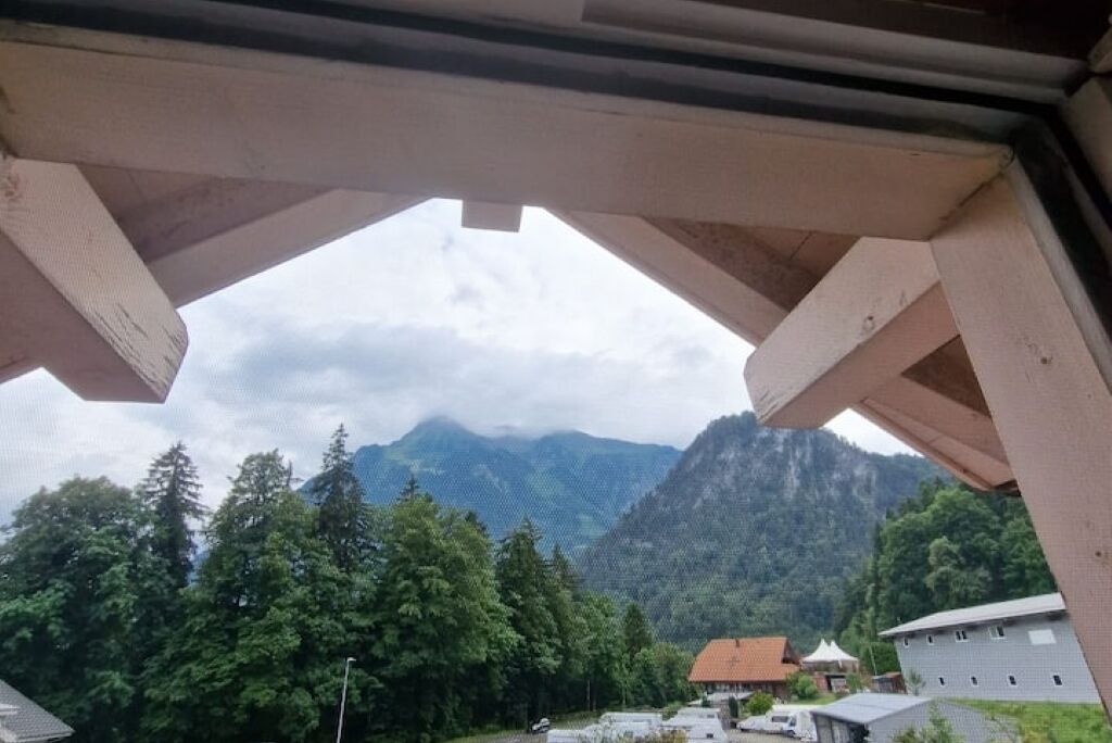 Vista a través de una estructura de madera, con montañas, un área boscosa y edificios al fondo.