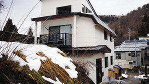 A modern two-story building with a balcony, surrounded by snow-covered vegetation. - Kogawa House  (Nozawaonsen)
