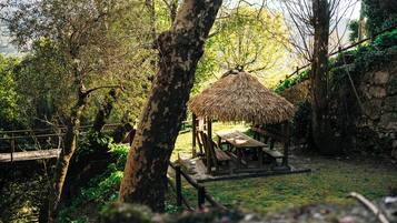 A rustic outdoor seating area with a thatched-roof structure, surrounded by trees and a stone wall.