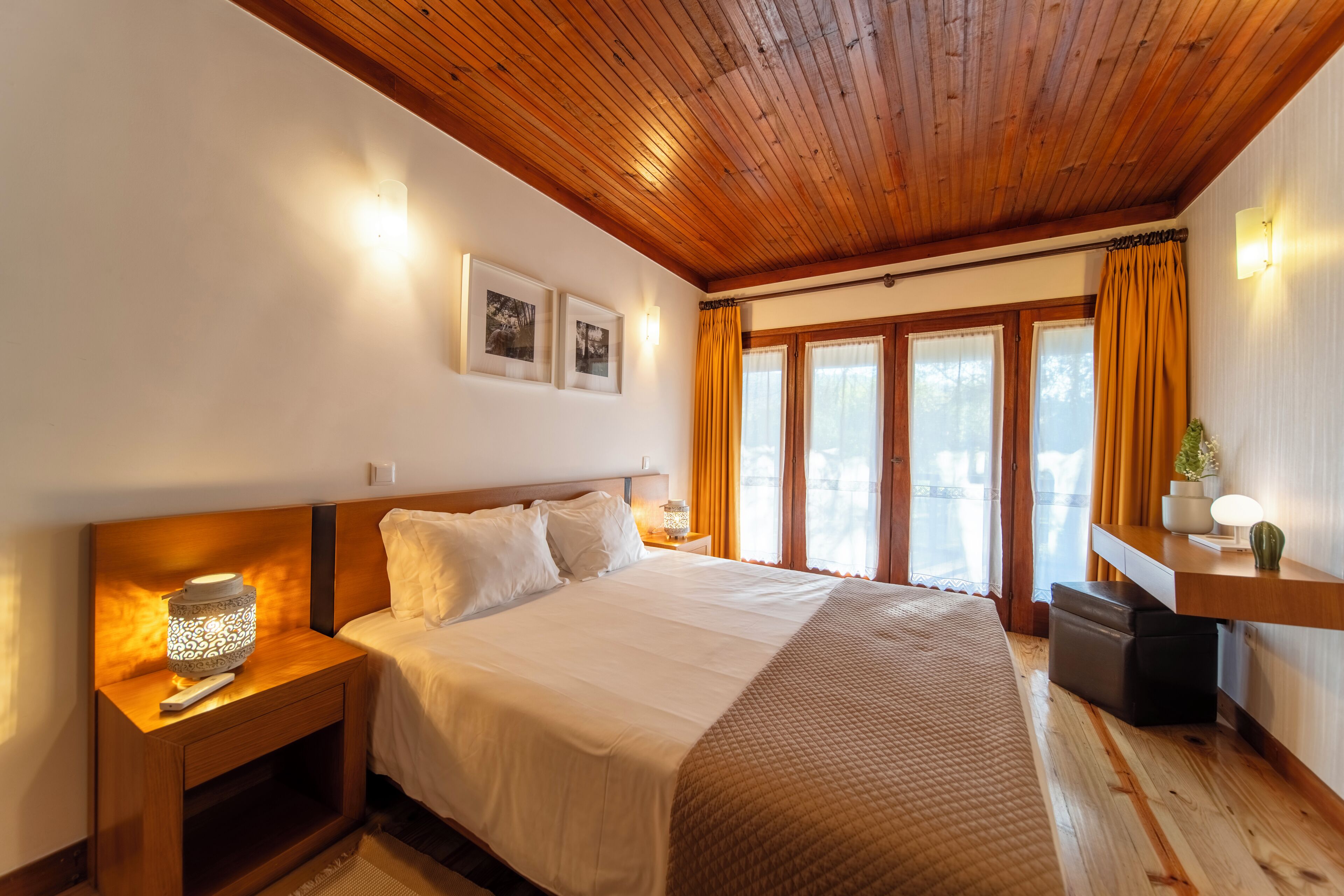 A hotel room with a wooden ceiling, a large bed with white bedding, a nightstand with a lamp, a small table with a plant, and a window with curtains.
