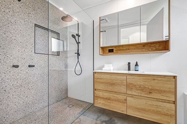 Un baño moderno con cabina de ducha de vidrio, tocador de madera con espejo y suelo de baldosas.