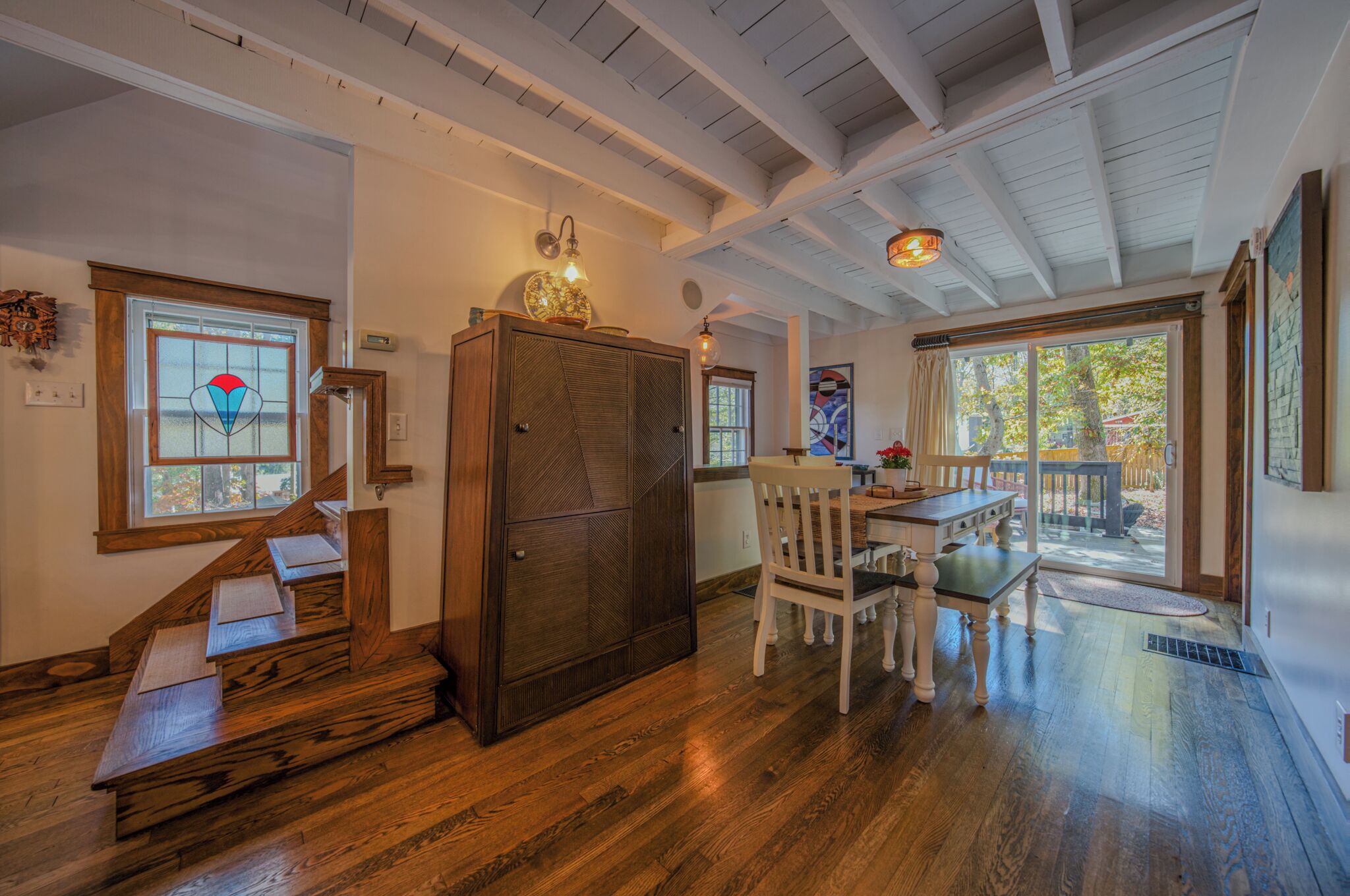 A wooden staircase leading to a dining area with a table and chairs, a large cabinet, and a view of the outdoors through a glass door.