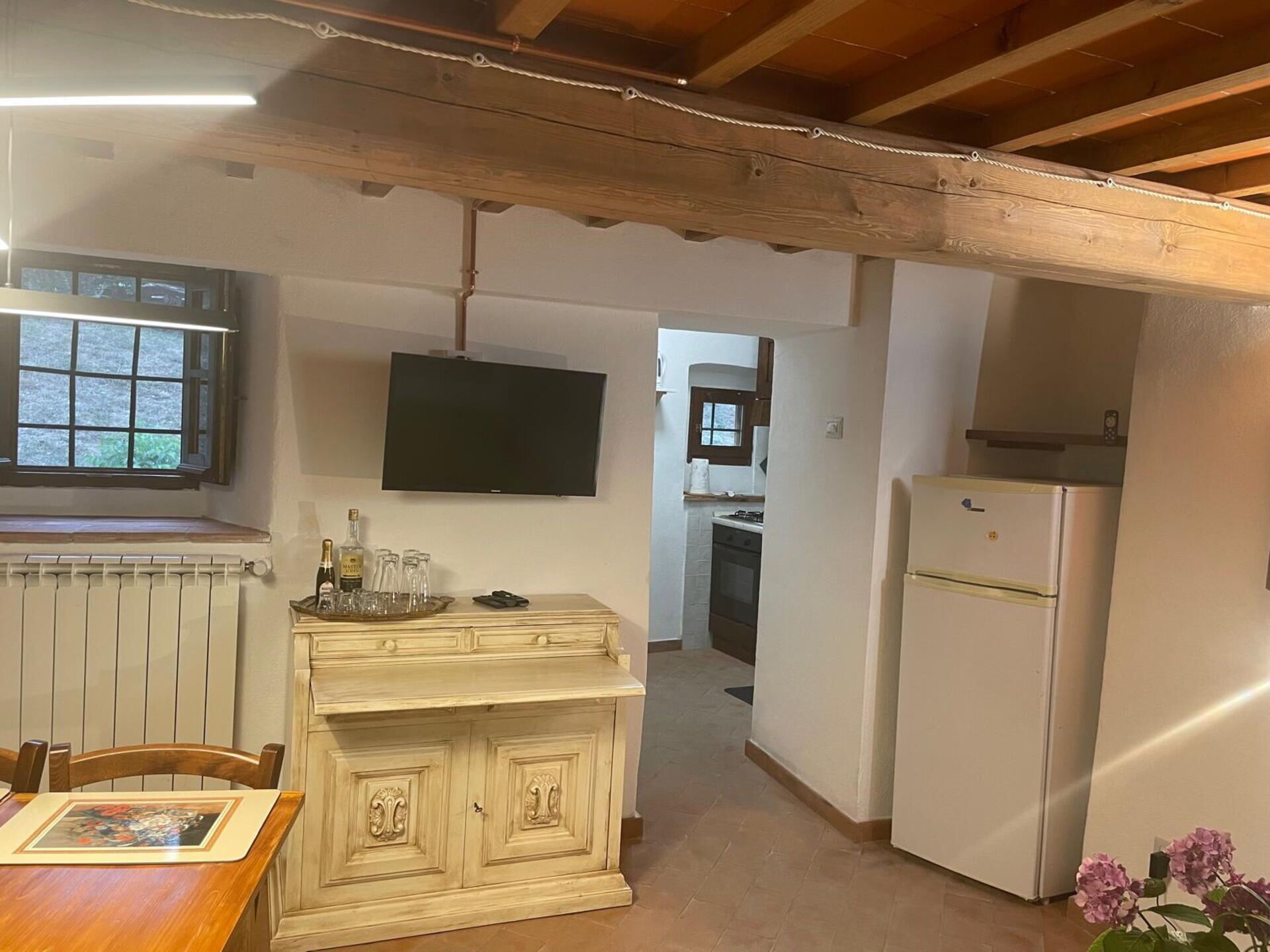 Una cocina con un refrigerador blanco, una mesa de comedor de madera con sillas, un televisor montado en la pared y un ventanal con vistas.