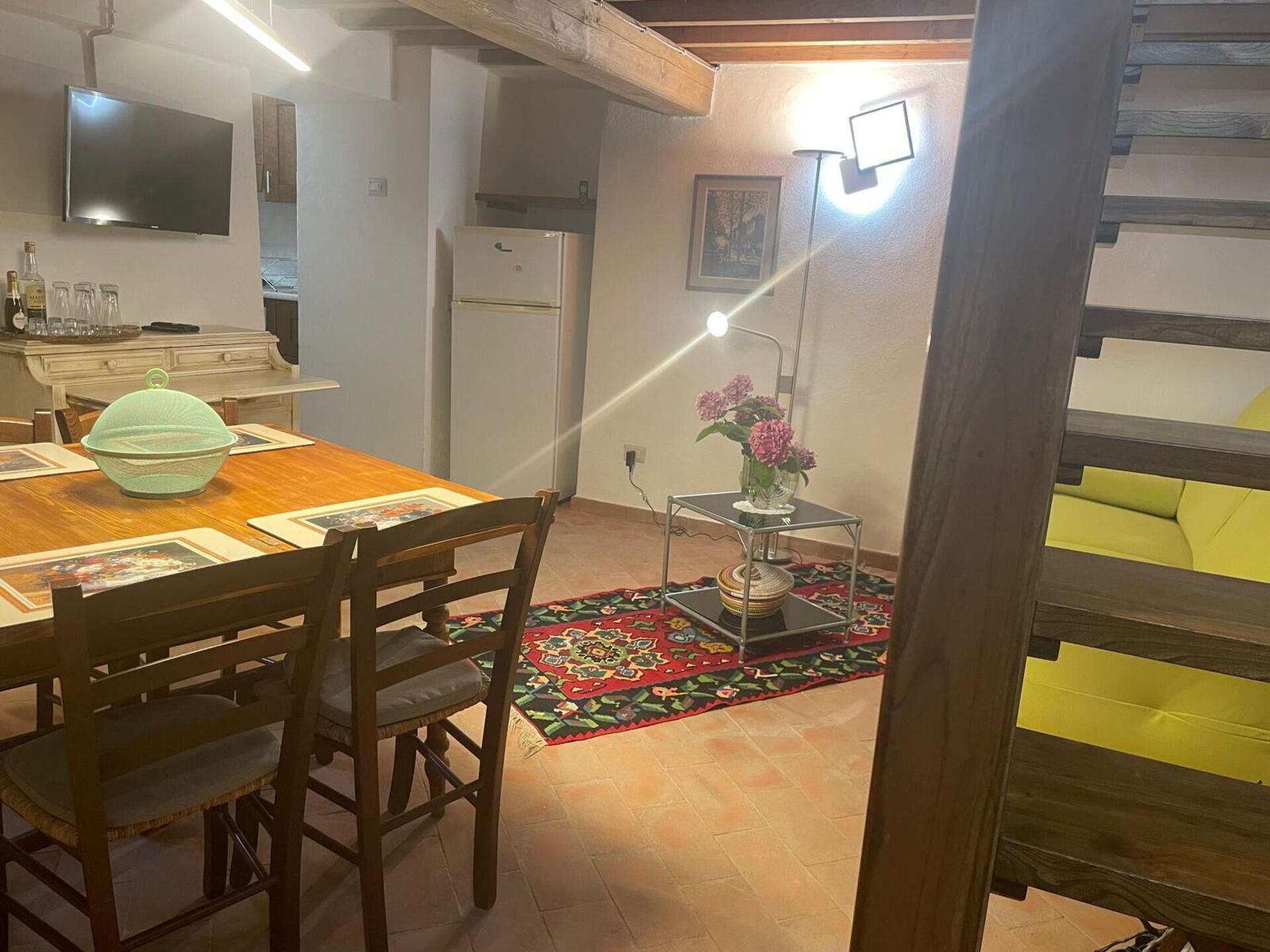A dining area with a wooden table, chairs, a decorative bowl, a refrigerator, a TV, a patterned rug, and a small table with a vase of flowers.