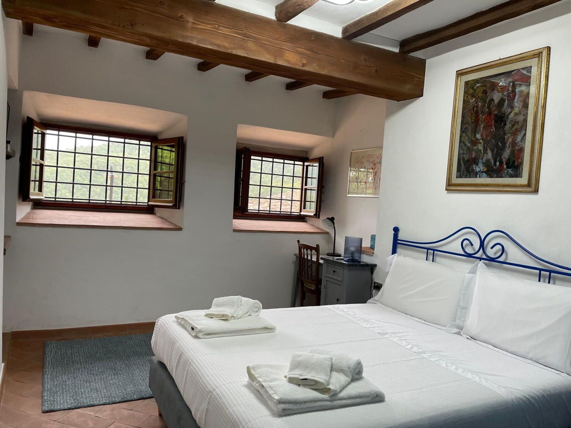 A bedroom with a bed, two windows, a desk, and a painting on the wall.