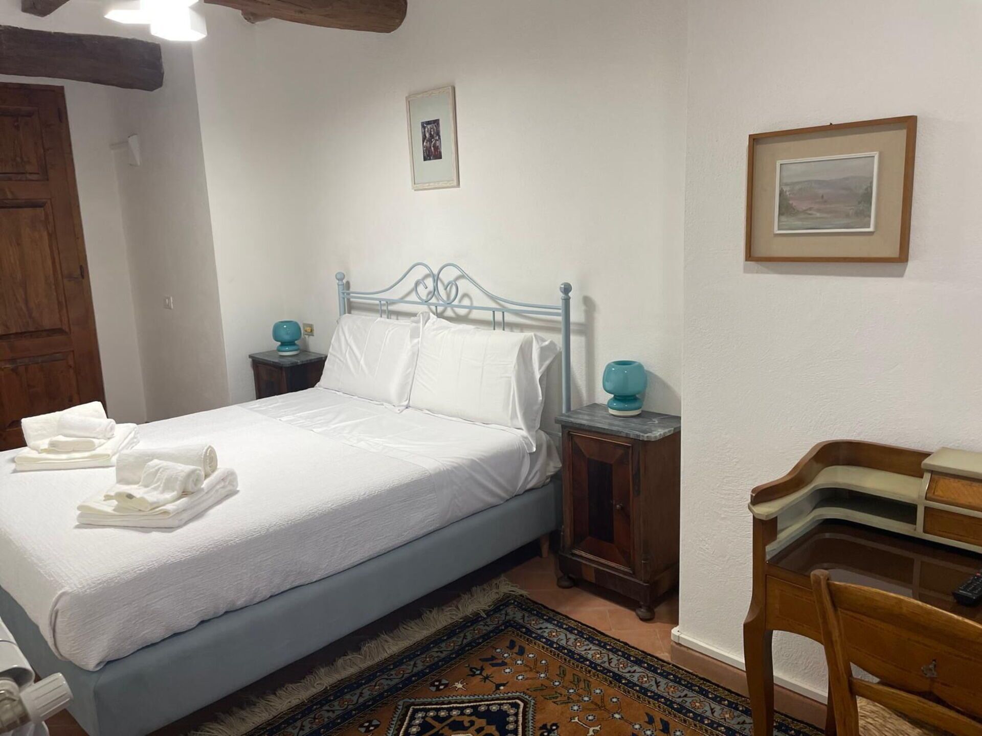 A bedroom with a bed, two bedside tables, a wooden chair, a desk, and a patterned rug.
