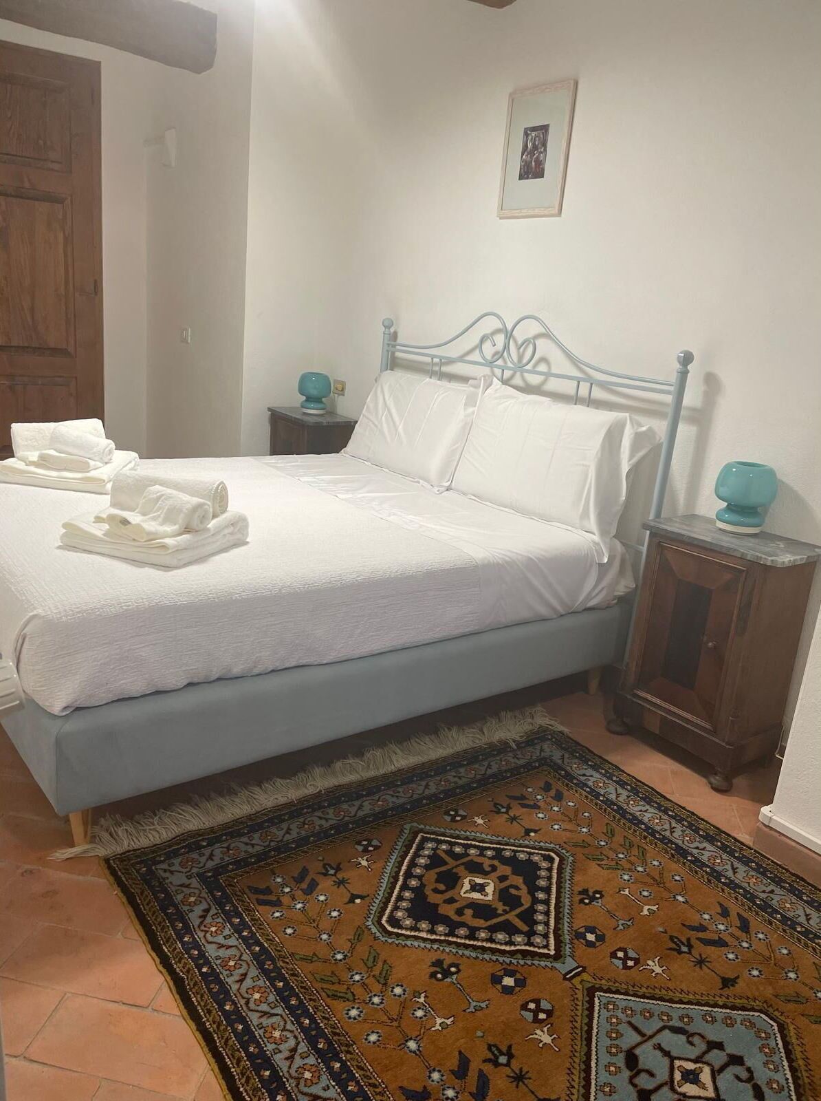 Un lit avec du linge de lit blanc, un tapis à motifs, une porte en bois et une petite table de chevet sur laquelle se trouve une lampe turquoise.