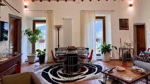 Een eethoek met een tafel, stoelen en een vloerkleed. Er staan potplanten, een lamp en een bank met kussens.