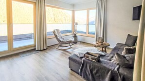 Un salon spacieux comprenant un canapé, un fauteuil à bascule, une petite table et de grandes fenêtres donnant sur l’extérieur.