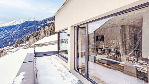 Un bâtiment moderne doté de grandes fenêtres en verre, d’un balcon enneigé et d’un paysage montagneux en arrière-plan.