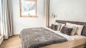 Quarto com cama de madeira, janela com vista para árvores, abajur e uma mesinha.