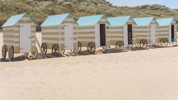 Una hilera de cabañas de playa con un diseño sencillo a rayas y techo plano, cada una con una puerta y una rueda a un lado.