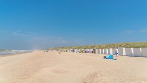 Una playa con una fila de cabañas de playa, una tienda de campaña y una colina cubierta de césped al fondo.