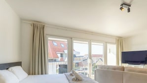 Habitación de hotel con cama, sofá, televisión y ventanas amplias con vista a edificios.