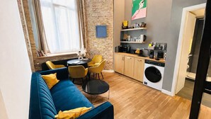 Un espace de vie compact avec un canapé bleu, des chaises jaunes, une petite table ronde, un coin cuisine équipé d’un lave-linge et un mur en briques.
