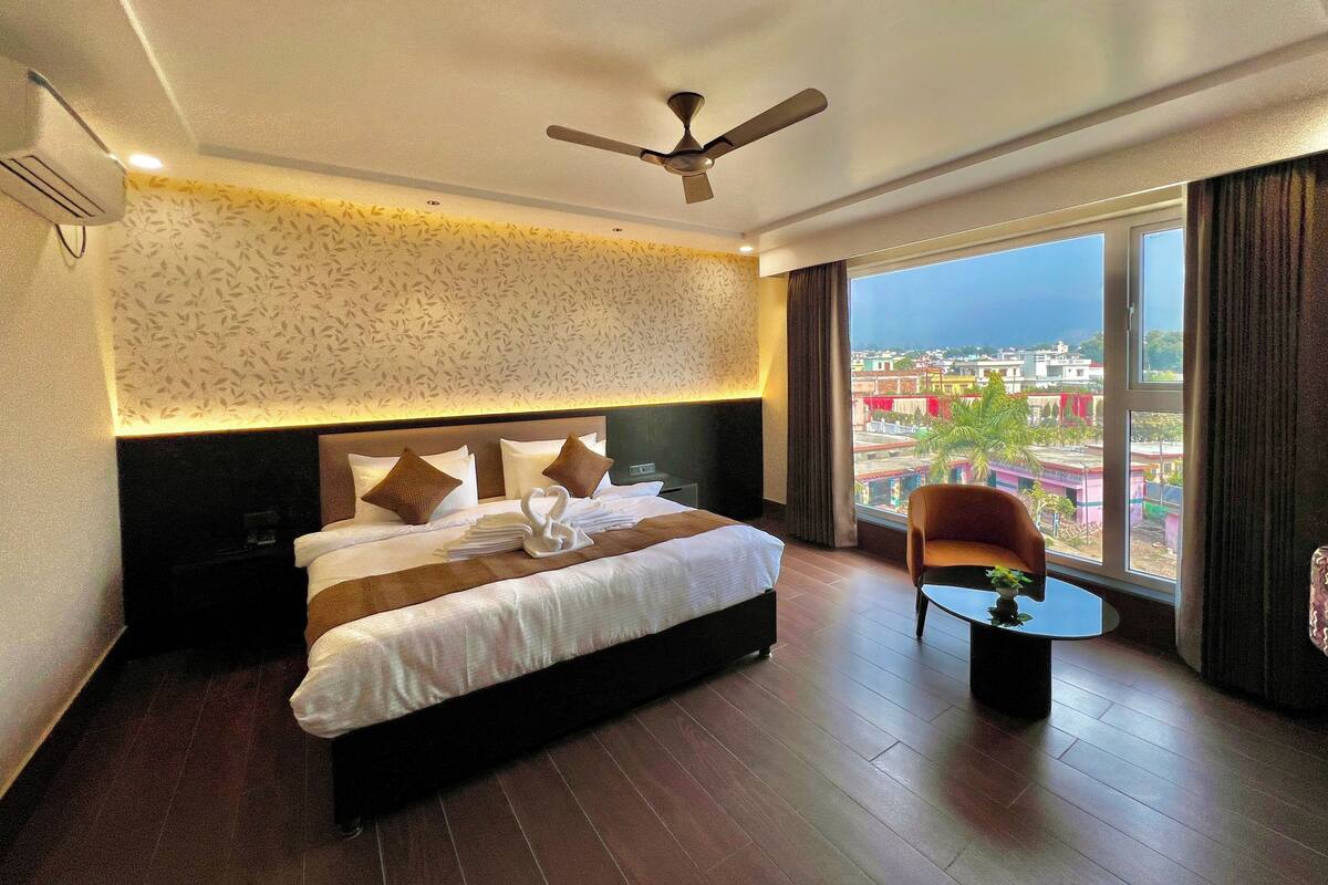 Habitación de hotel con una cama grande, ventilador de techo, vista a la ciudad y una pequeña mesa con una planta.