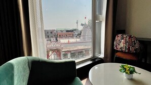 A room with a green chair, a small table with a plant, and a window with a view of buildings and a flag.