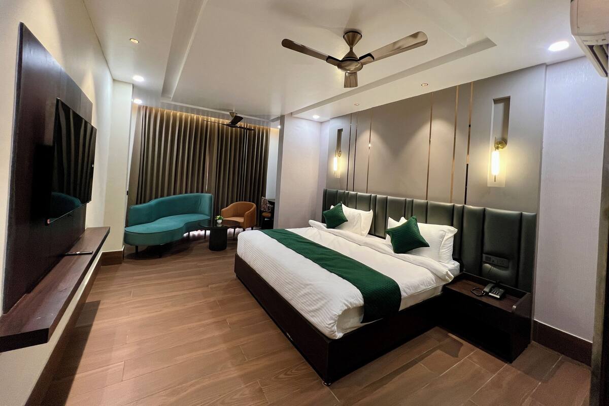 Una habitación de hotel moderna con cama, televisor, ventilador de techo, zona de estar con sofá y sillón, y televisor montado en la pared.