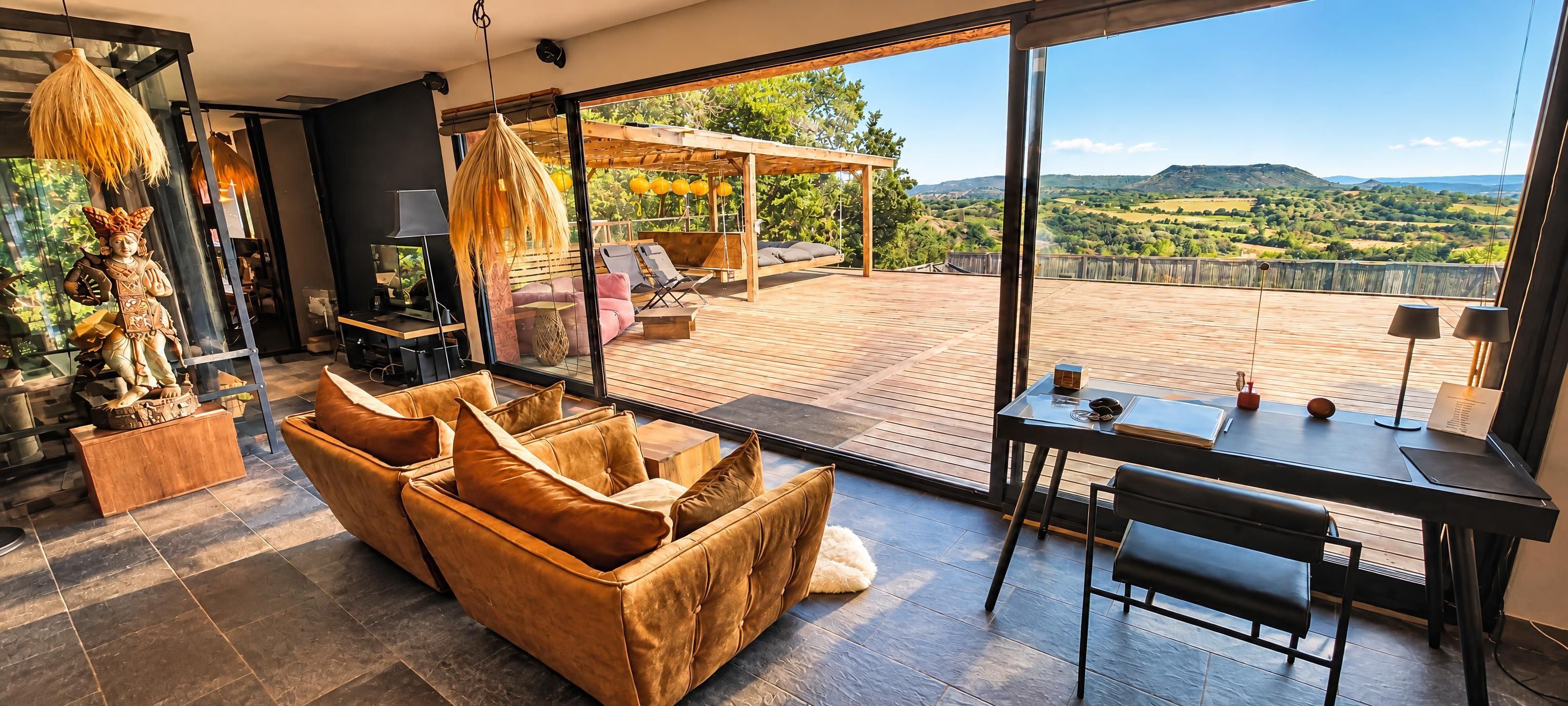 A modern living room with a large window, a sofa, a dining table, and a view of a scenic landscape.