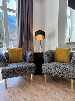 Ein modernes Wohnzimmer mit zwei gemusterten Sesseln, einer schwarzen Stehlampe und Blick auf Berge durch große Fenster.