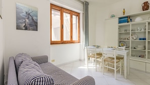 Un salon avec un canapé à rayures, une table à manger avec des chaises, un coin cuisine avec des étagères et une fenêtre avec des rideaux.