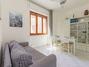 Ein Wohnzimmer mit einem gestreiften Sofa, einem Esstisch mit Stühlen, einer Küchenzeile mit Regalen und einem Fenster mit Vorhängen.