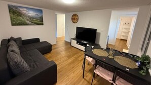 A living room with a sectional sofa, a flat-screen TV, a dining table with chairs, and a painting on the wall. - Happy Life 2-20min Interlaken-Panoramic view of mountains (Wimmis)