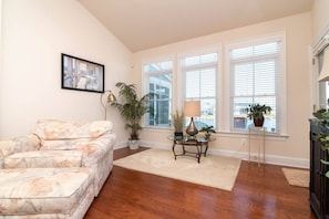 A living room with a sofa, a coffee table, and a lamp. There are three windows with blinds, and a plant is on the windowsill. - Waterfront Resort | Community Amenities | Sleeps 13 (Selbyville)