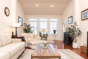 A living room with a sofa, armchair, coffee table, and a fireplace. There are windows with blinds, a clock on the wall, and a plant. - Waterfront Resort | Community Amenities | Sleeps 13 (Selbyville)