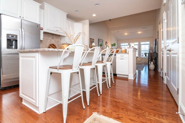 A modern kitchen with white cabinetry, a stainless steel refrigerator, a built-in island with bar stools, and a wooden floor. - Waterfront Resort | Community Amenities | Sleeps 13 (Selbyville)