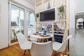 A modern living room with a glass door leading to a balcony, a mounted TV, and a dining area with white chairs and a table set with blue and white dishes. - Charming studio apartment with a view of the sea in Novi Vinodolski (Novi Vinodolski)