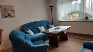 Een woonkamer met een blauwe bank, een houten bijzettafeltje, een vloerlamp en een raam met uitzicht op groen.