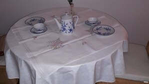 Een ronde tafel met een wit tafelkleed, een karaf, kopjes en schoteltjes.