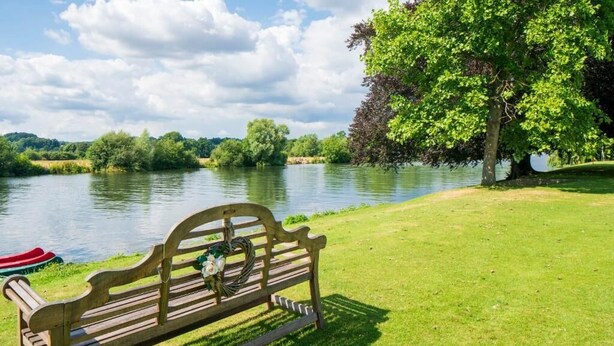 A wooden bench with a wreath, positioned on a grassy bank beside a calm river. - Bisham Abbey National Sports Centre  (Bisham)