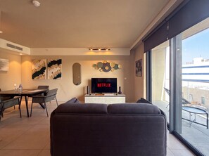 A modern living room with a flat-screen TV displaying 'NETFLIX,' a dining area with a table and chairs, and a balcony with a view of the city. - 6th Floor Mercury Suite Apartment with Pool Access (Saint Julian's)
