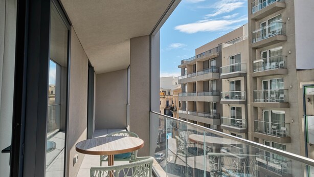 Een balkon met een tafel, stoelen, een glazen hekwerk en uitzicht op omliggende gebouwen.