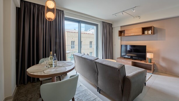 Et moderne hotelværelse med sofa, fjernsyn, spisebord med stole og et stort vindue med gardiner.