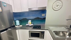 A compact kitchen with a refrigerator, microwave, sink, and a scenic wall mural. - Coastal Breeze Nelson Bay: King Bed, Spa bath, Private balcony, Resort pool (Nelson Bay)
