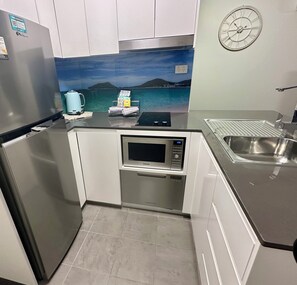 A compact kitchen with a refrigerator, microwave, sink, and a scenic wall mural. - Coastal Breeze Nelson Bay: King Bed, Spa bath, Private balcony, Resort pool (Nelson Bay)