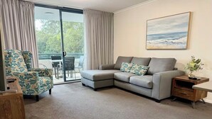 A living room with a grey sofa, a floral armchair, a wooden side table, and a view through a sliding glass door to an outdoor area. - Coastal Breeze Nelson Bay: King Bed, Spa bath, Private balcony, Resort pool (Nelson Bay)