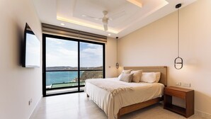Une chambre à coucher comprenant un lit, une table de chevet, un ventilateur de plafond et une grande fenêtre donnant sur la mer.
