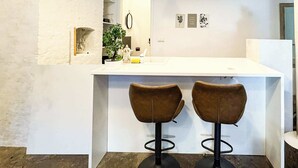 Eine Küche mit einer weißen Kücheninsel, zwei braunen Barhocker, Pendelleuchten und einem Blumentopf.