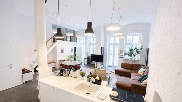 Eine moderne Küche mit Kochinsel, ein Essbereich mit rundem Tisch, ein Wohnzimmer mit braunem Sofa und eine Treppe, die zu einer oberen Etage führt.